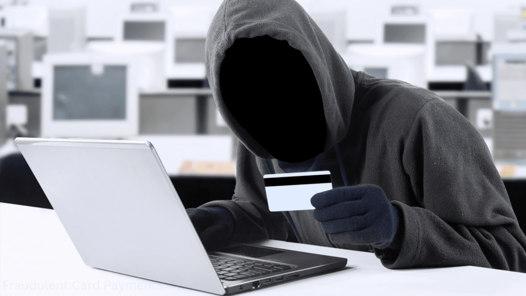 A faceless figure in a hoodie and gloves holds a credit card in front of a laptop, suggesting cybercrime in a room with blurred computer equipment.